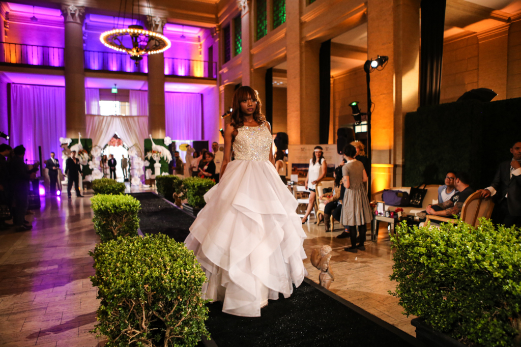 Bride model on runway inside venue for vanity wedding show with purple wall lights in the background and chandelier lighting