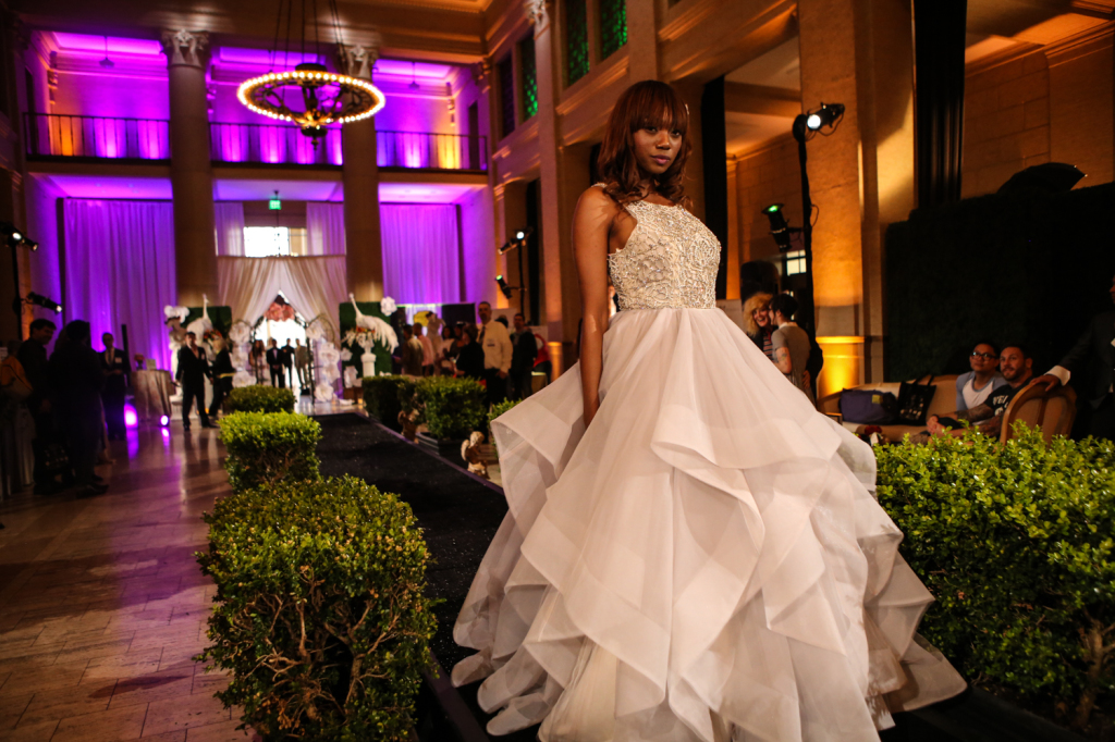 Bride model on runway for vanity wedding show with purple lighting on back wall and chandelier lighting