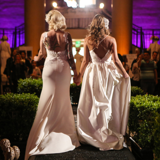 Two bride models on runway underneath chandelier lights for gay vanity wedding show