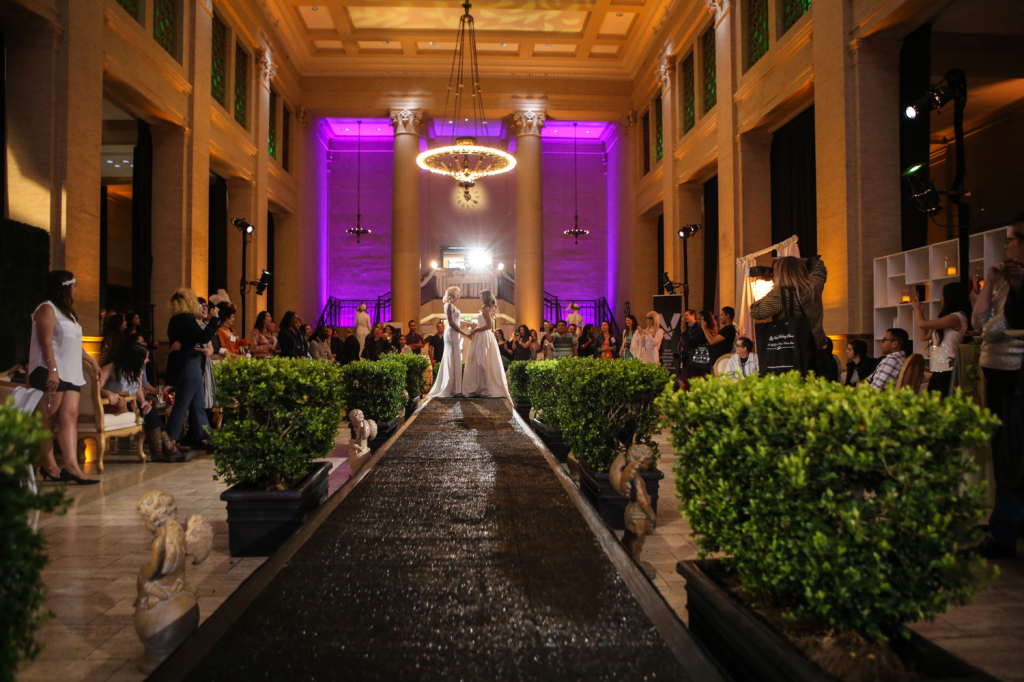 Two bride models on runway posing for gay vanity wedding show underneath chandelier lights 