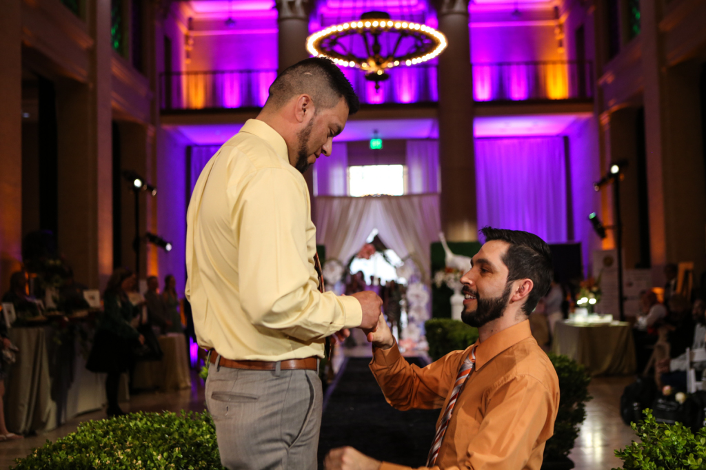 Proposal on runway underneath chandelier lights for gay vanity wedding show and purple lights wall in the background