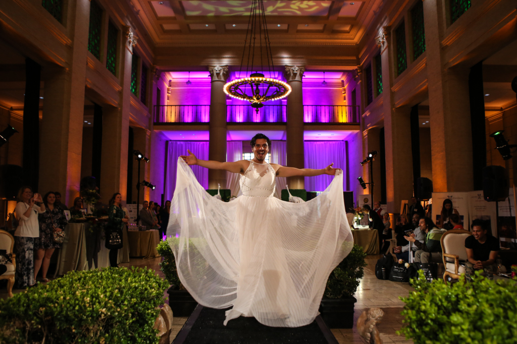 Bride model posing on runway for gay vanity wedding show with chandelier lights and purple wall lights in the background