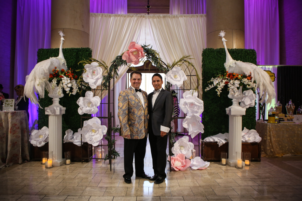 Two men posing in front of flower and statue installation with drapery and purple wall lights in the background for gay vanity wedding show